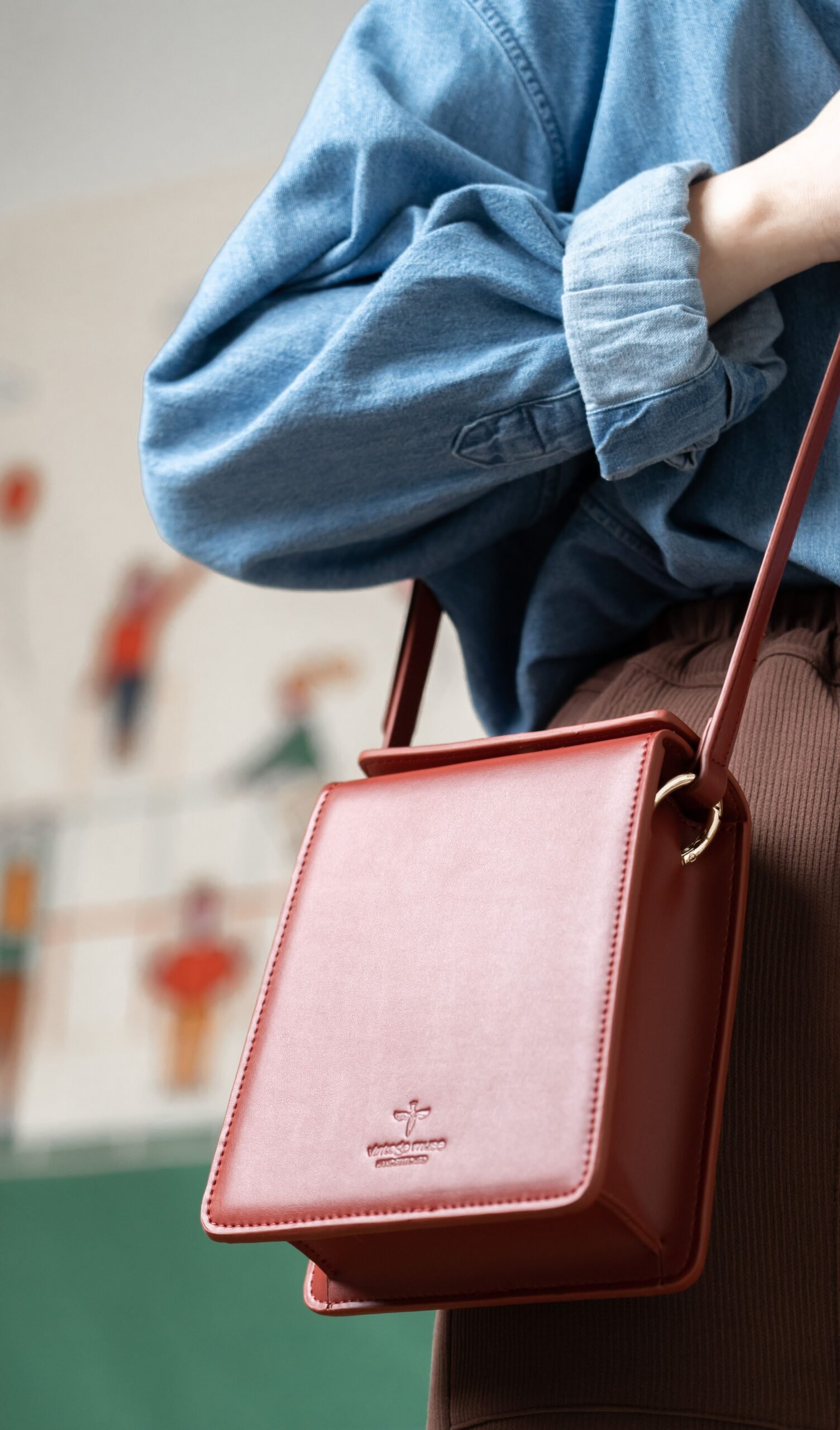 Close-up of a stylish red leather sling bag worn over a rolled-up denim shirt, indoors.